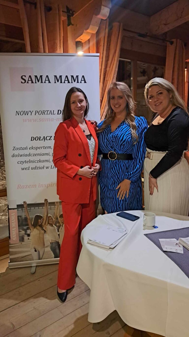 Trzy kobiety przed plakatem Fundacji Sama Mama. Jedna w pomarańczowym kostiumie, druga w niebieskiej sukience w czarne paski , a trzecia w białej spódnicy i czarnej bluzce