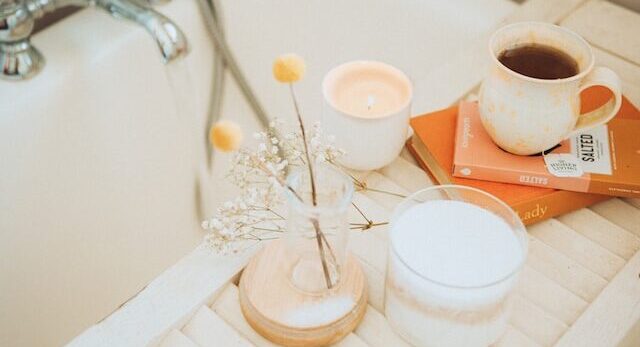 Biała wanna ze stolikiem, na którym znajduje się wazonik z kwiatkami książka, kubek z kawą i zapaloną świeczką. Opis pokazuje przykład przyjemnego rytuału