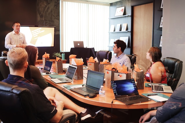 Grupa ludzi na szkoleniu, wspomagająca się laptopami, siedzi przy stole i słucha prowadzącego, który stoi przed tablicą